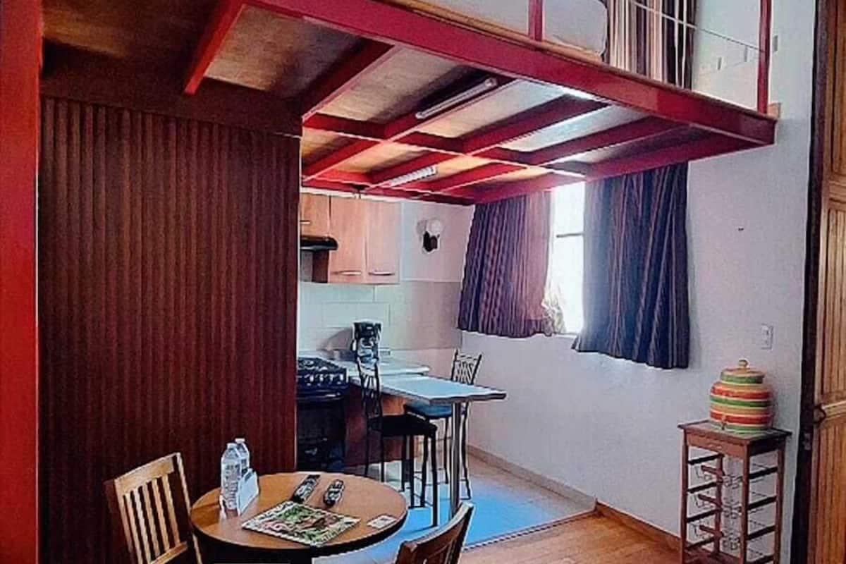 A dining area with a wooden table and chairs, a red accent wall, and a view of a kitchen and upper level.