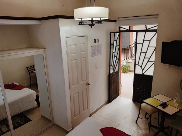 Habitación pequeña con cama, escritorio y silla. Se ve una puerta que da a un área exterior con barandilla.