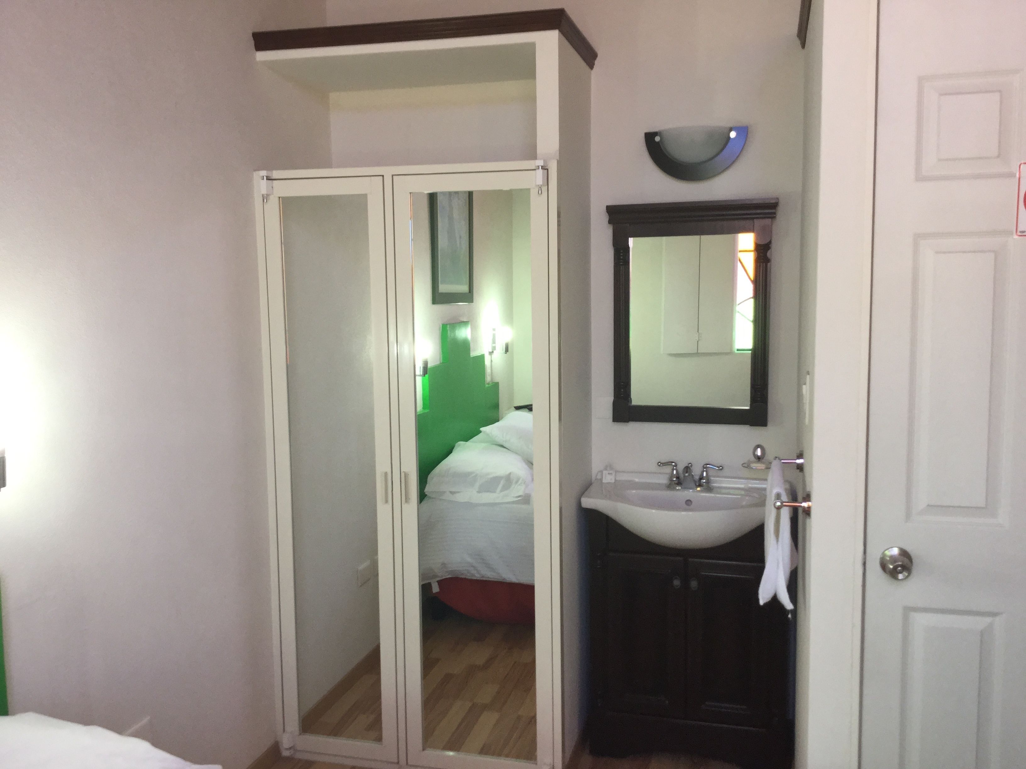 Habitación de hotel con vestidor, baño con lavamanos y espejo, y cama con cabecera verde.