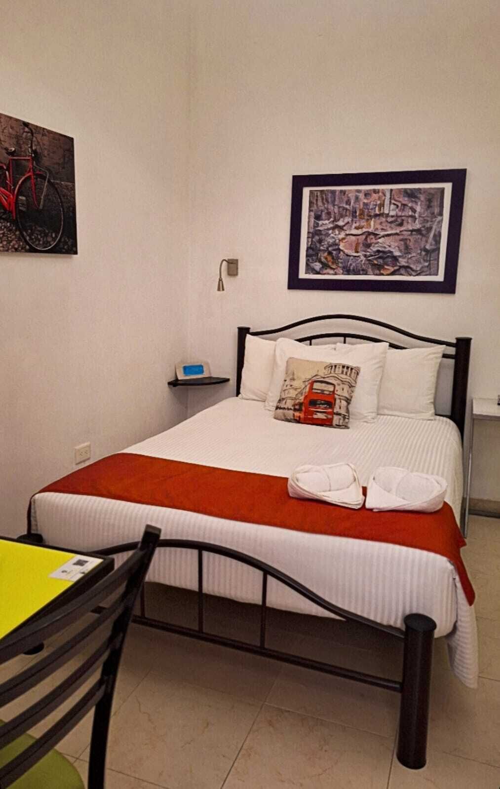 A neatly made bed with white bedding and a red blanket, a small bedside table with a lamp, and a chair with a yellow table.