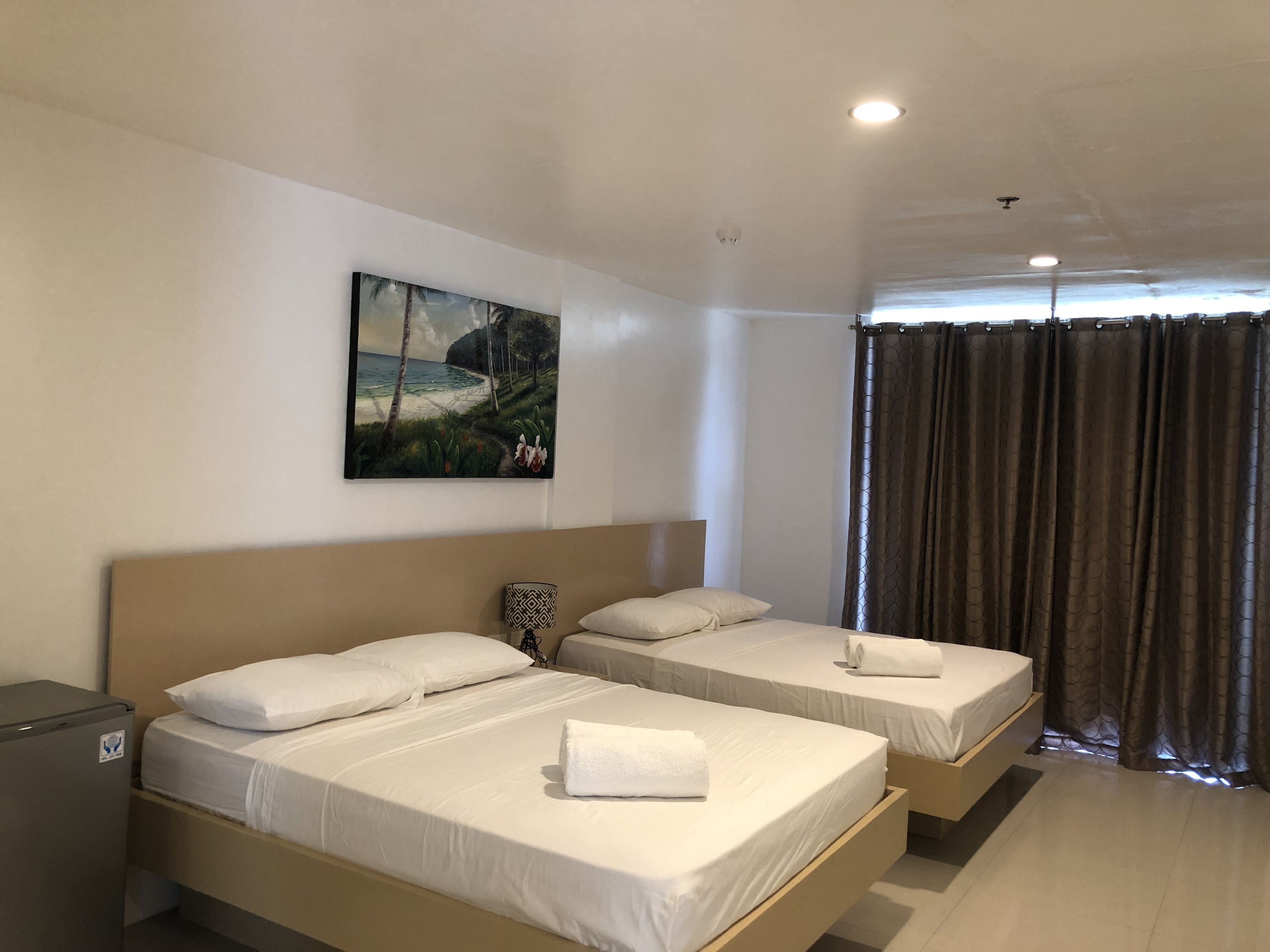 Een hotelkamer met twee eenpersoonsbedden, een klein bureau, een koelkast, een schilderij van een strandgezicht en een raam met gordijnen.