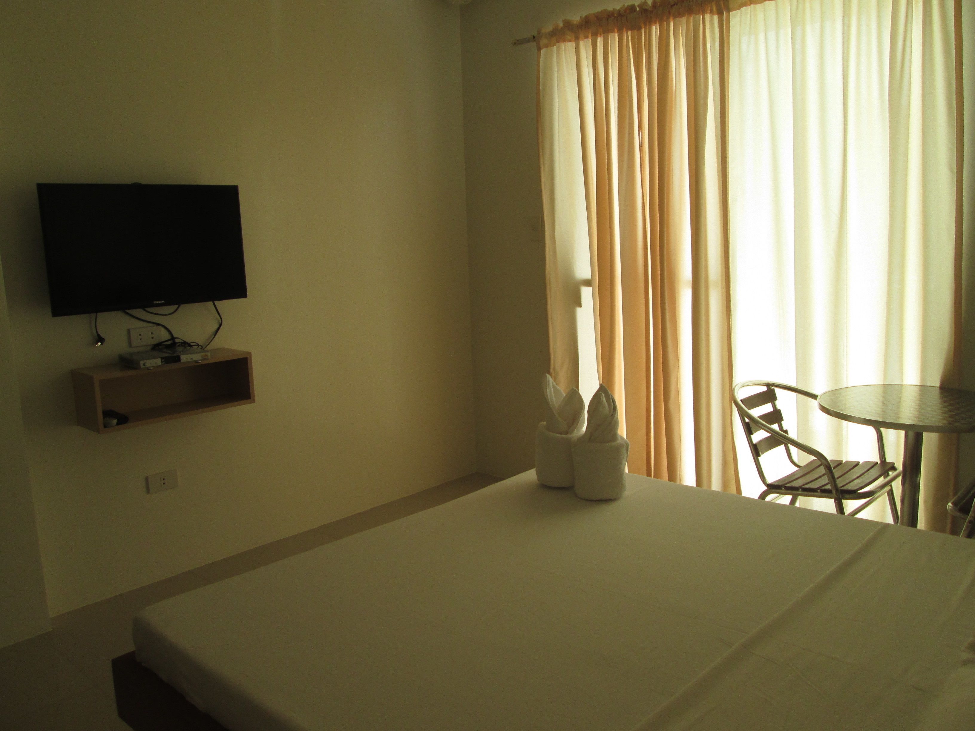 Habitación de hotel con cama, televisión montada en la pared, una mesita con una silla y una ventana con cortinas.