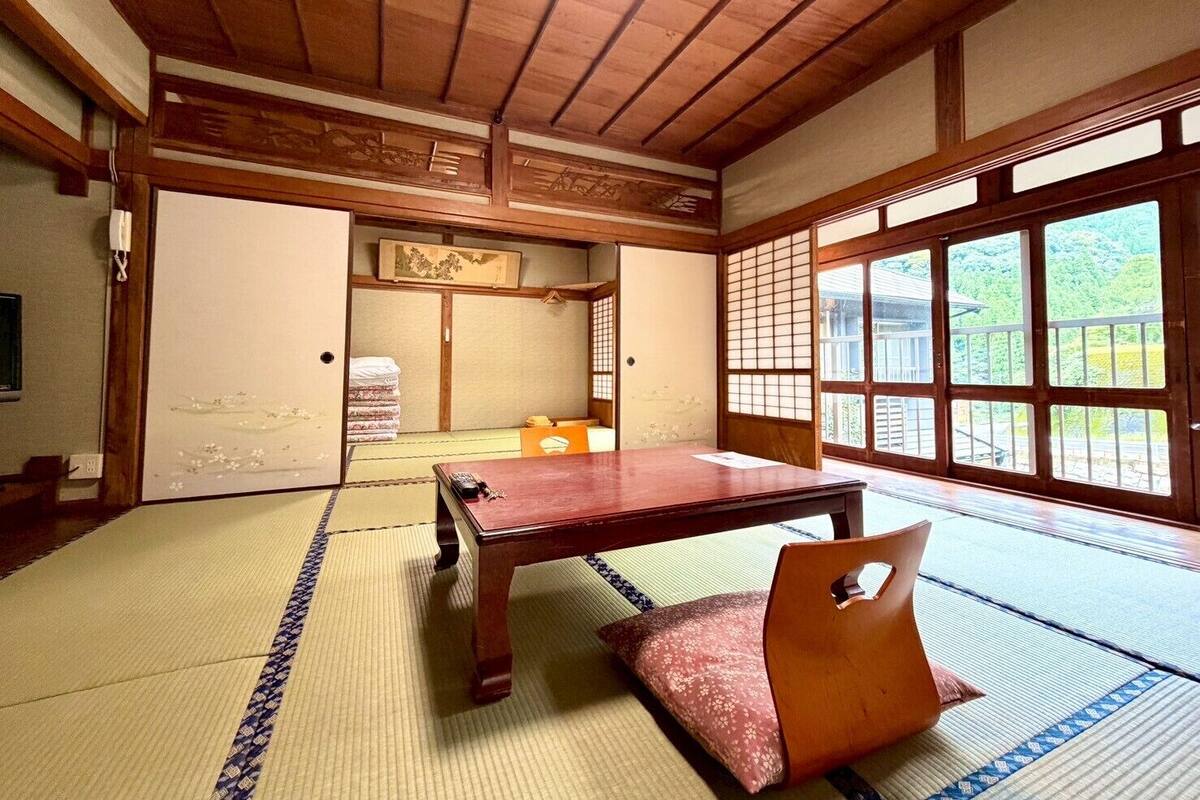 Una habitación japonesa tradicional con suelo de tatami, una mesa de madera baja y un fusuma que da a un balcón con vistas a la vegetación.