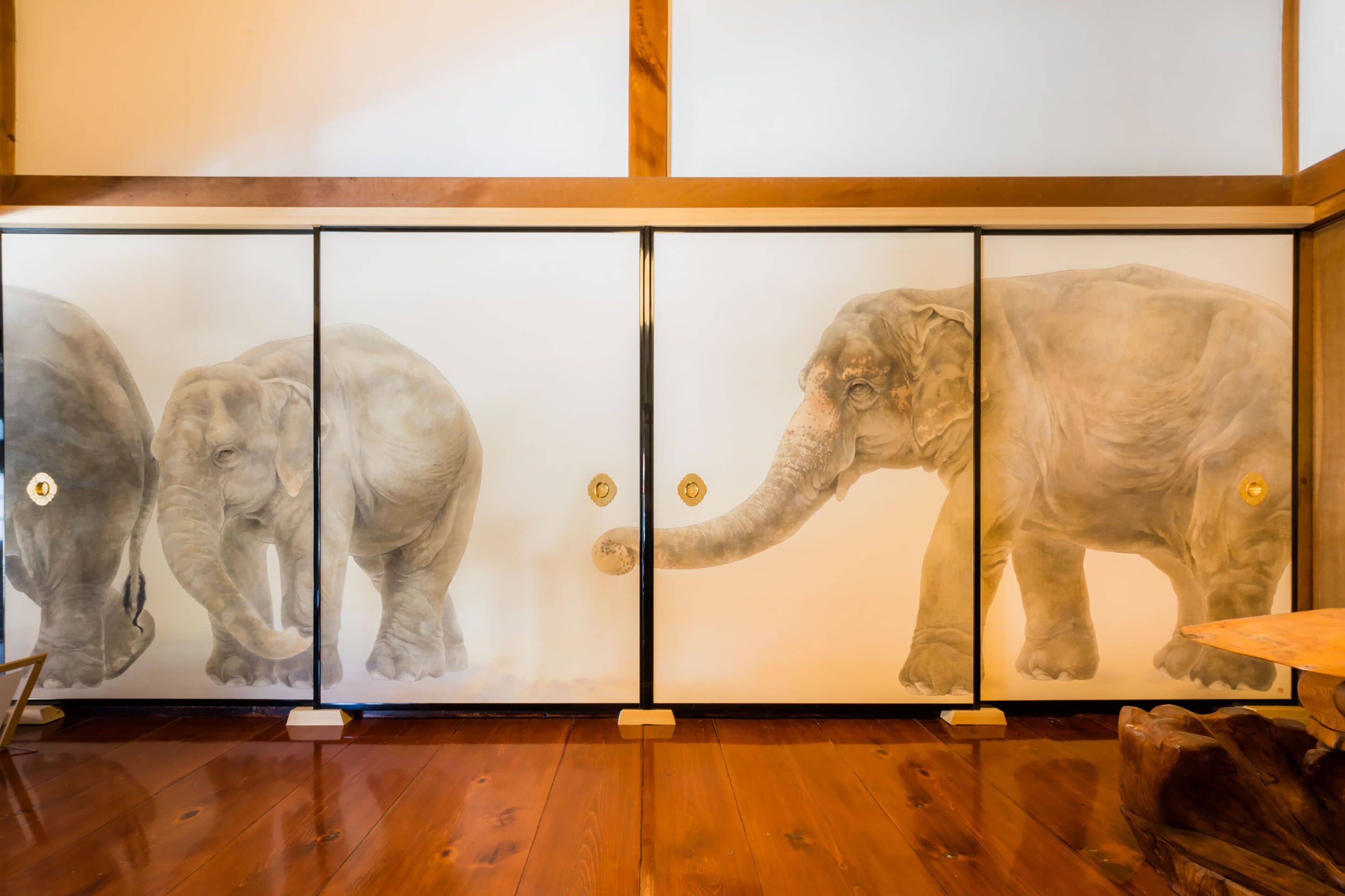 A room with a wooden floor, a wooden table, and a cabinet with glass doors displaying elephant artwork.