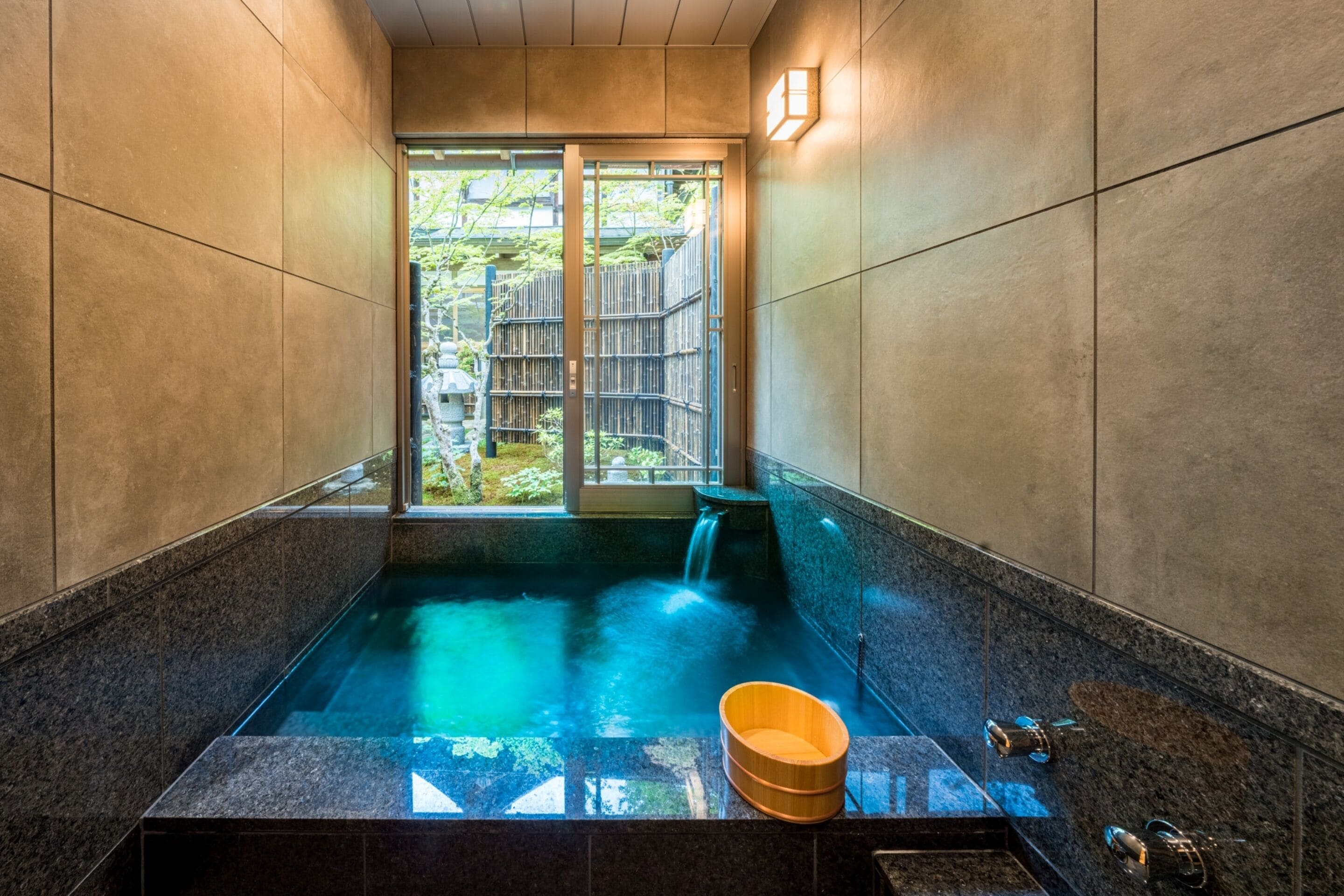 A modern spa with a large hot tub, a wooden bucket, and a view of a garden through a glass door.