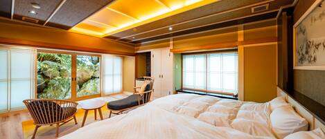 A spacious bedroom with a large bed, a small table, and a chair. There are large windows with a view of greenery outside.
