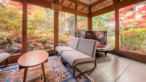 A traditional Japanese-style room with tatami flooring, a low wooden table, a cushioned bench, and large windows offering a view of a garden with autumn foliage.
