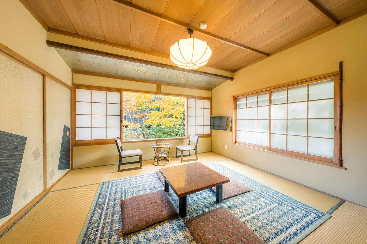 A traditional Japanese room with tatami flooring, a low table, cushions, and a view of autumn foliage through shoji screens.
