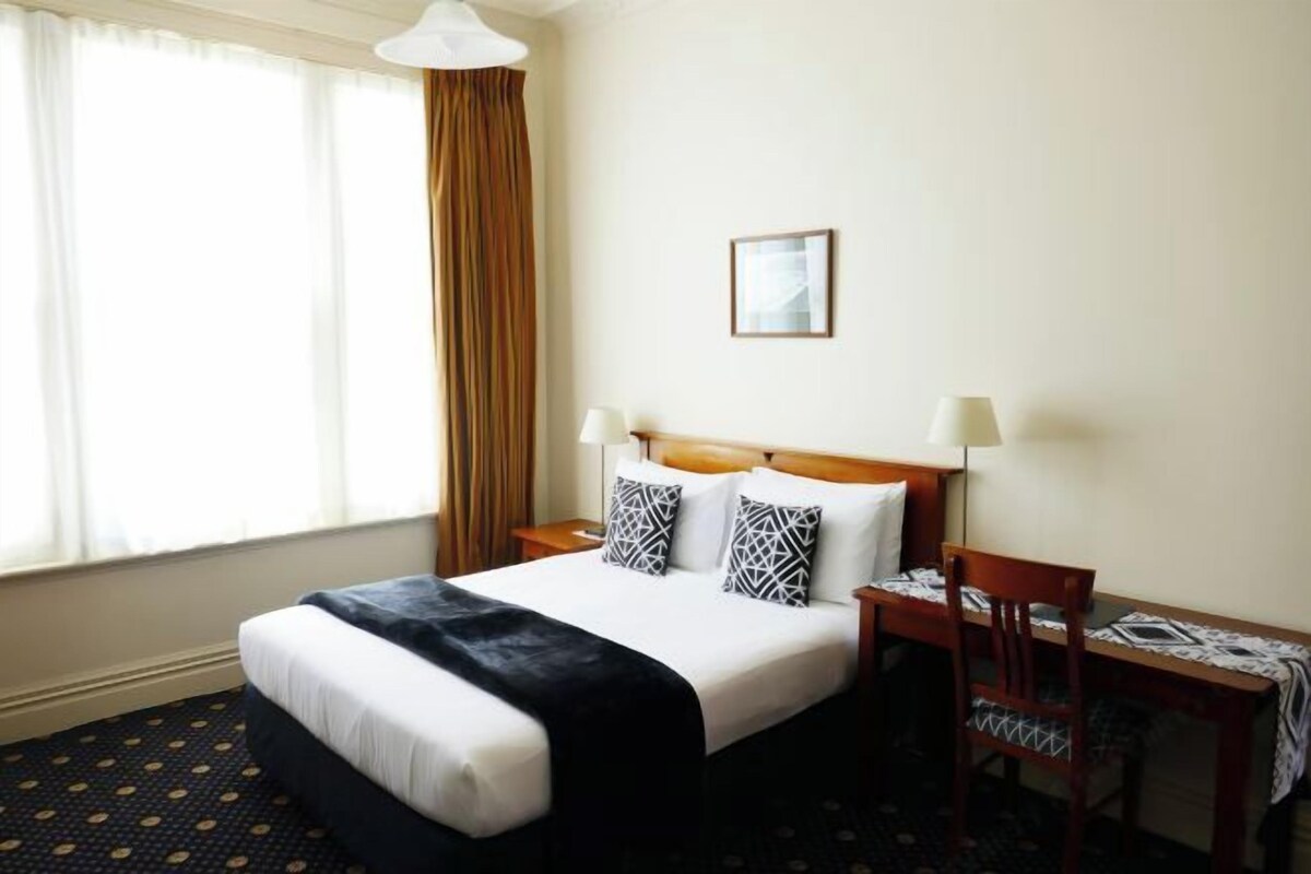 A hotel room with a bed, two bedside tables with lamps, a small table with a chair, and a window with curtains.