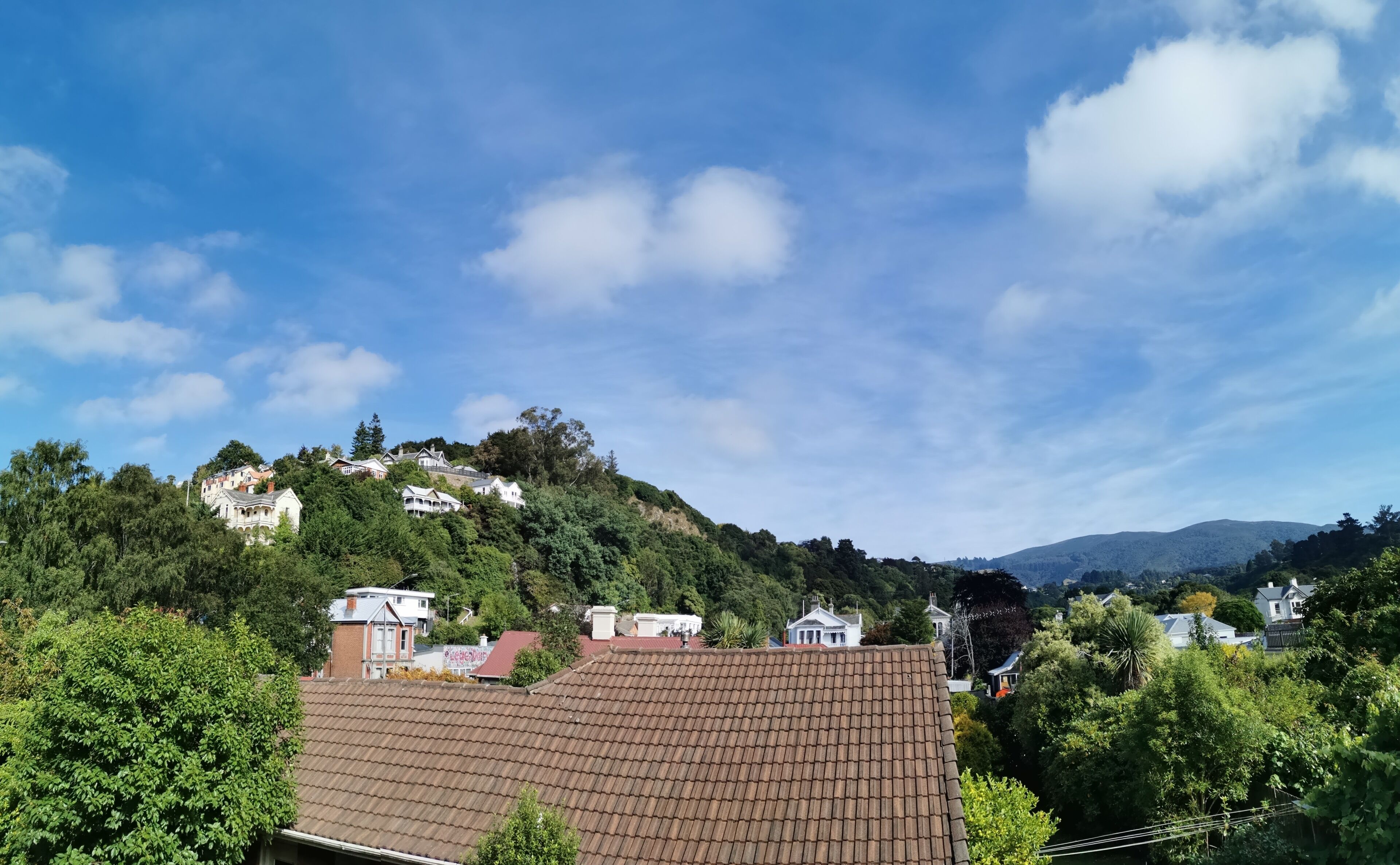 A residential area with houses on a hillside, surrounded by greenery and under a clear sky.