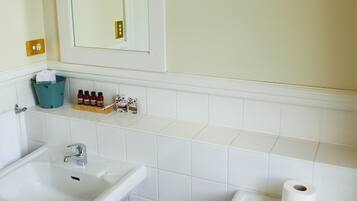 A bathroom with a white sink, toilet, and mirror. There is a trash can and a shelf with toiletries.