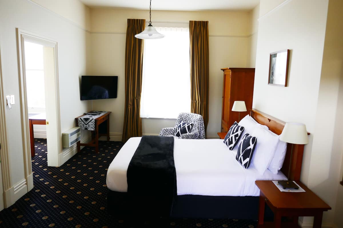Una habitación de hotel con cama, televisor, escritorio, silla, lámpara y ventana con cortinas.