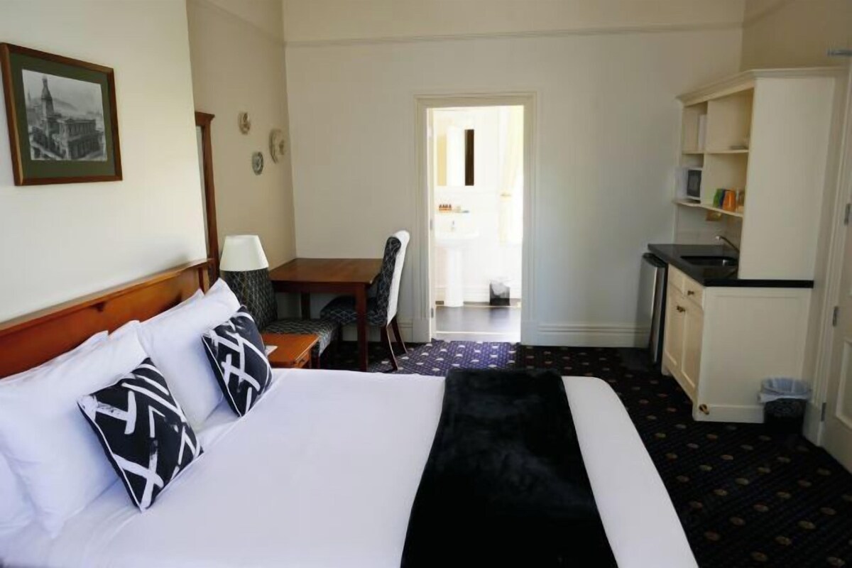 A hotel room with a bed, a desk, a chair, a small kitchenette, and a door leading to a bathroom.