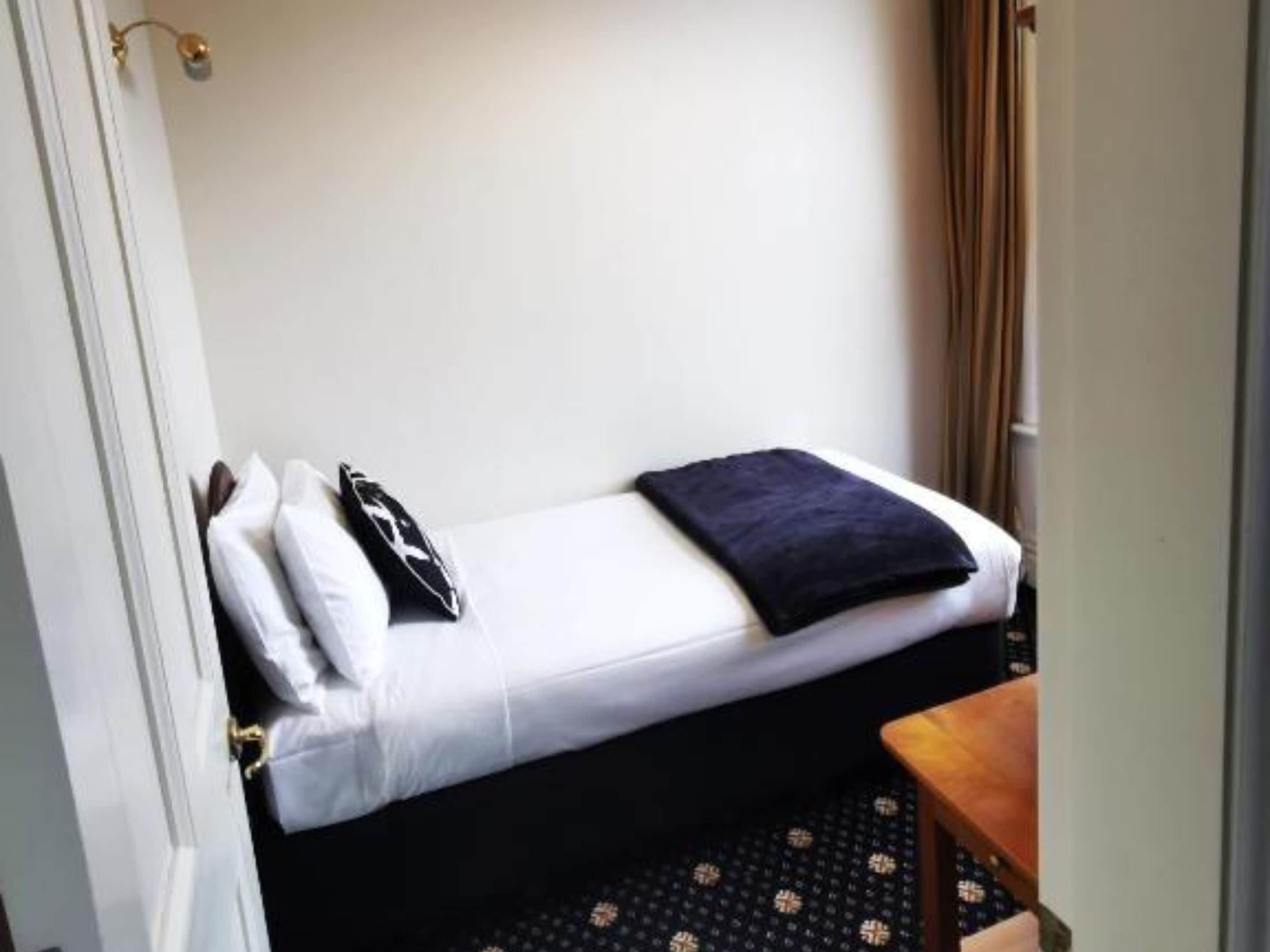 A small, single-bed room with a white bed, a dark blanket, and a black pillow.