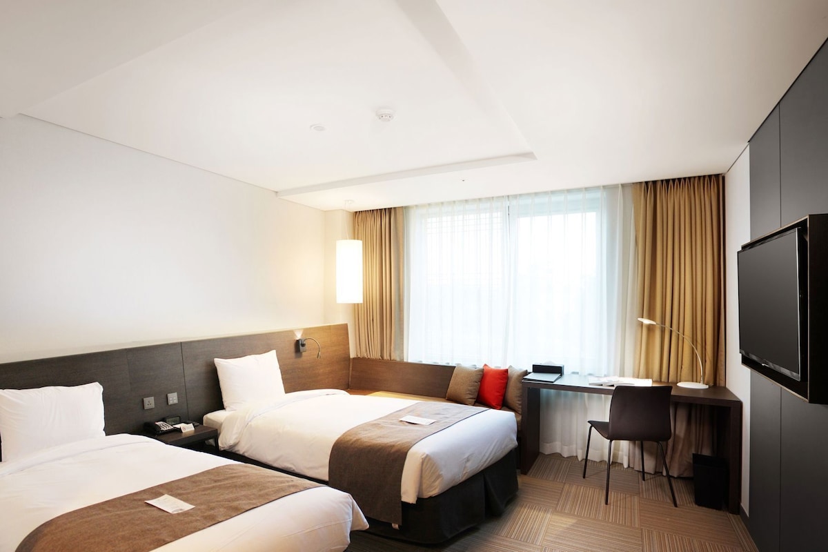 Une chambre d’hôtel avec deux lits, un bureau, une chaise, une télévision et une fenêtre avec des rideaux.