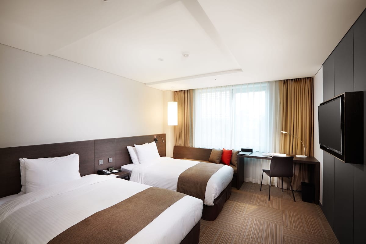 Une chambre d’hôtel avec deux lits, un bureau, une chaise, une télévision et une fenêtre avec des rideaux.