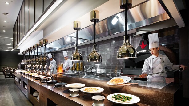 A modern kitchen with chefs preparing food, stainless steel appliances, and a variety of dishes displayed on the counter. - Hotel Prumir (Hwaseong)