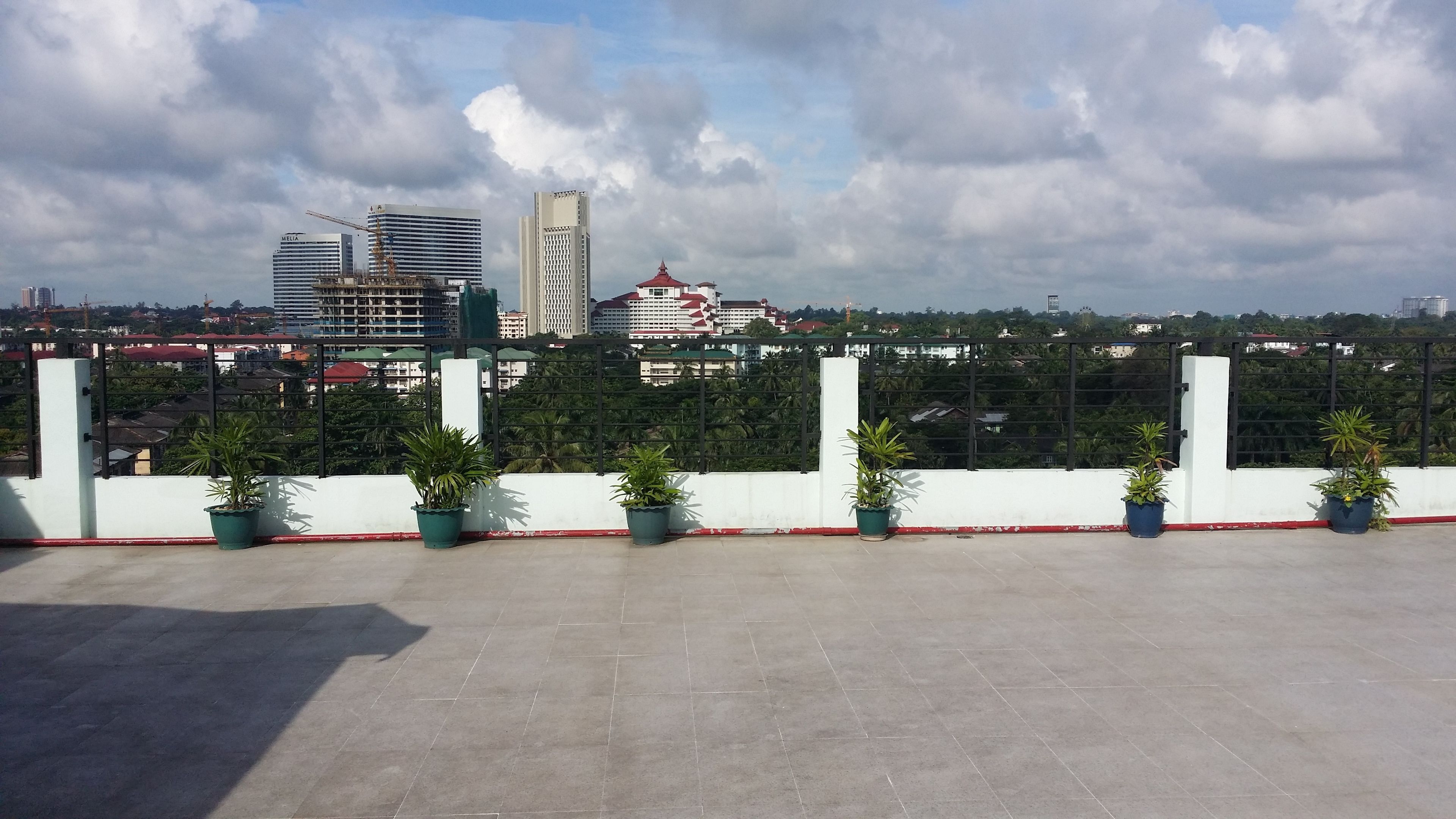 A rooftop terrace with potted plants, a white railing, and a view of city buildings in the background.
