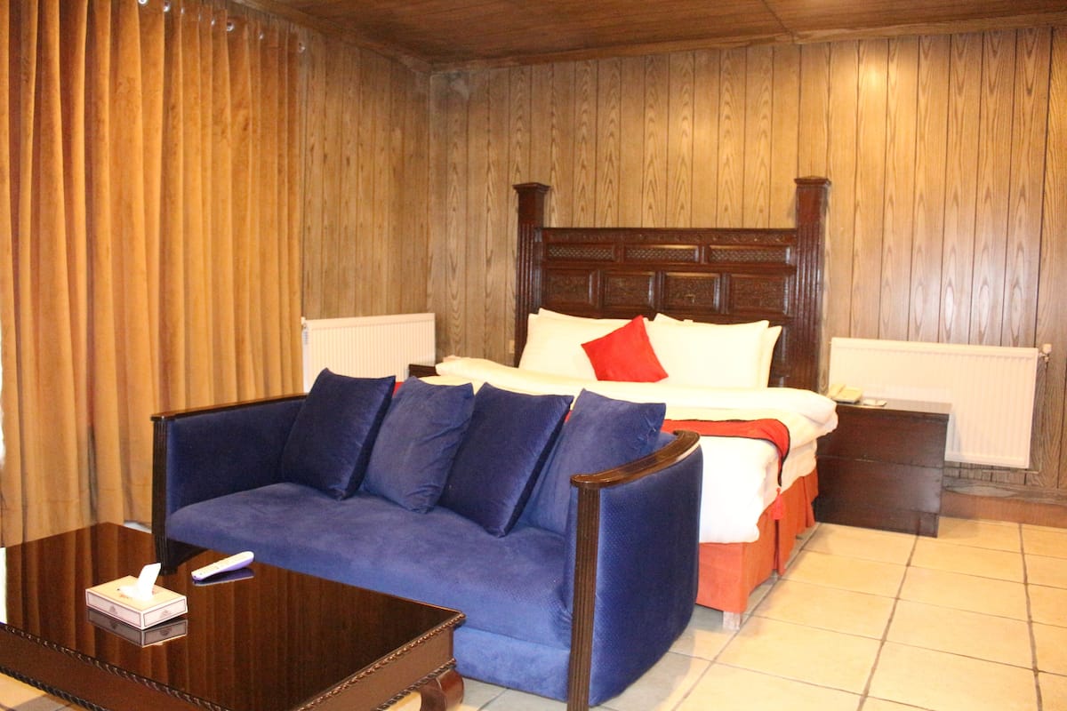 Un dormitorio con cama de madera con cabecera, un sofá azul, una mesita de noche de madera y una mesita auxiliar con lámpara.