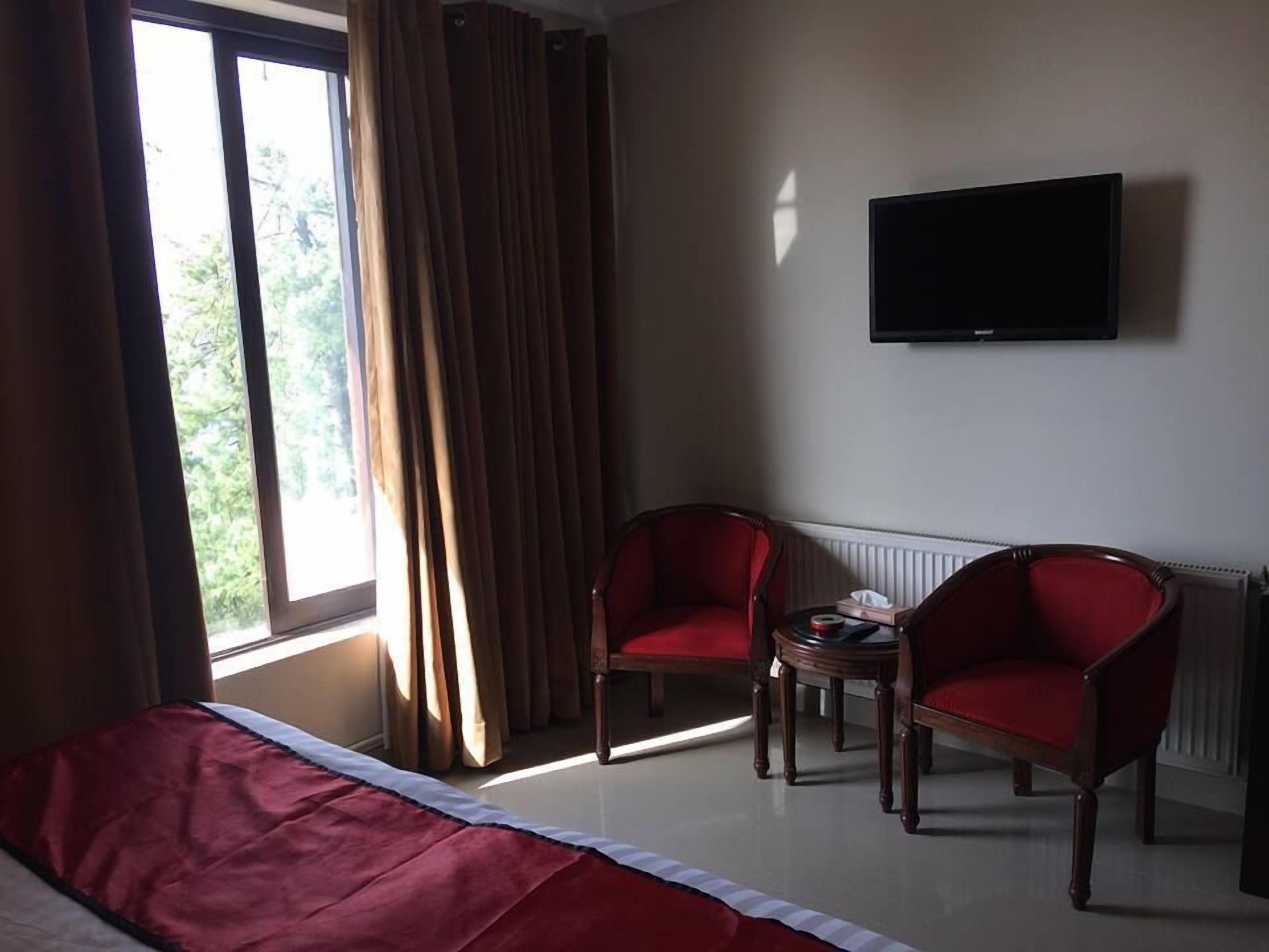 Quarto de hotel com uma cama, duas poltronas vermelhas, uma mesinha, uma televisão de tela plana na parede e uma janela com cortinas.