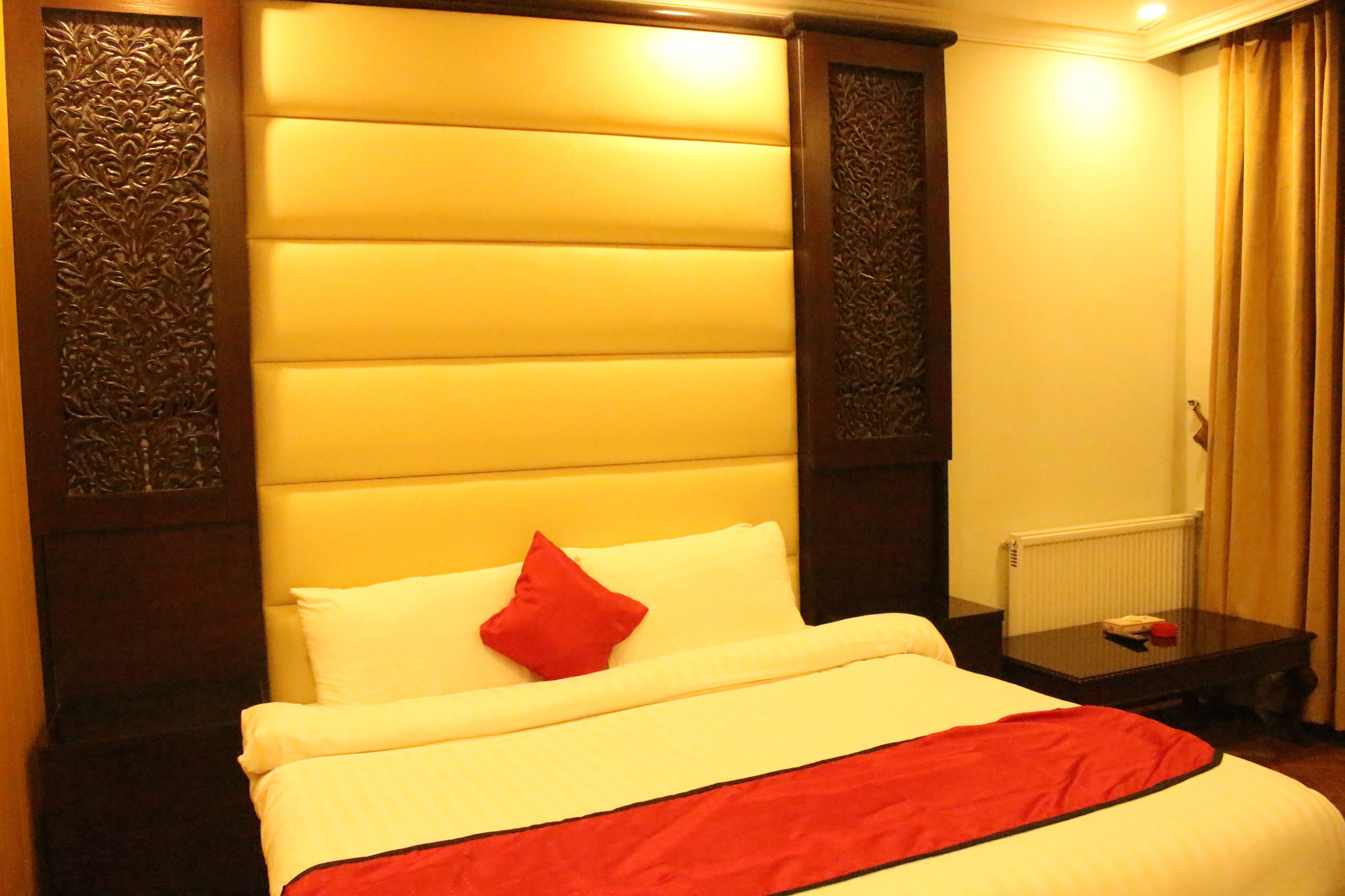 Una habitación de hotel con una cama grande, un cojín rojo, una mesita de noche de madera y un radiador.
