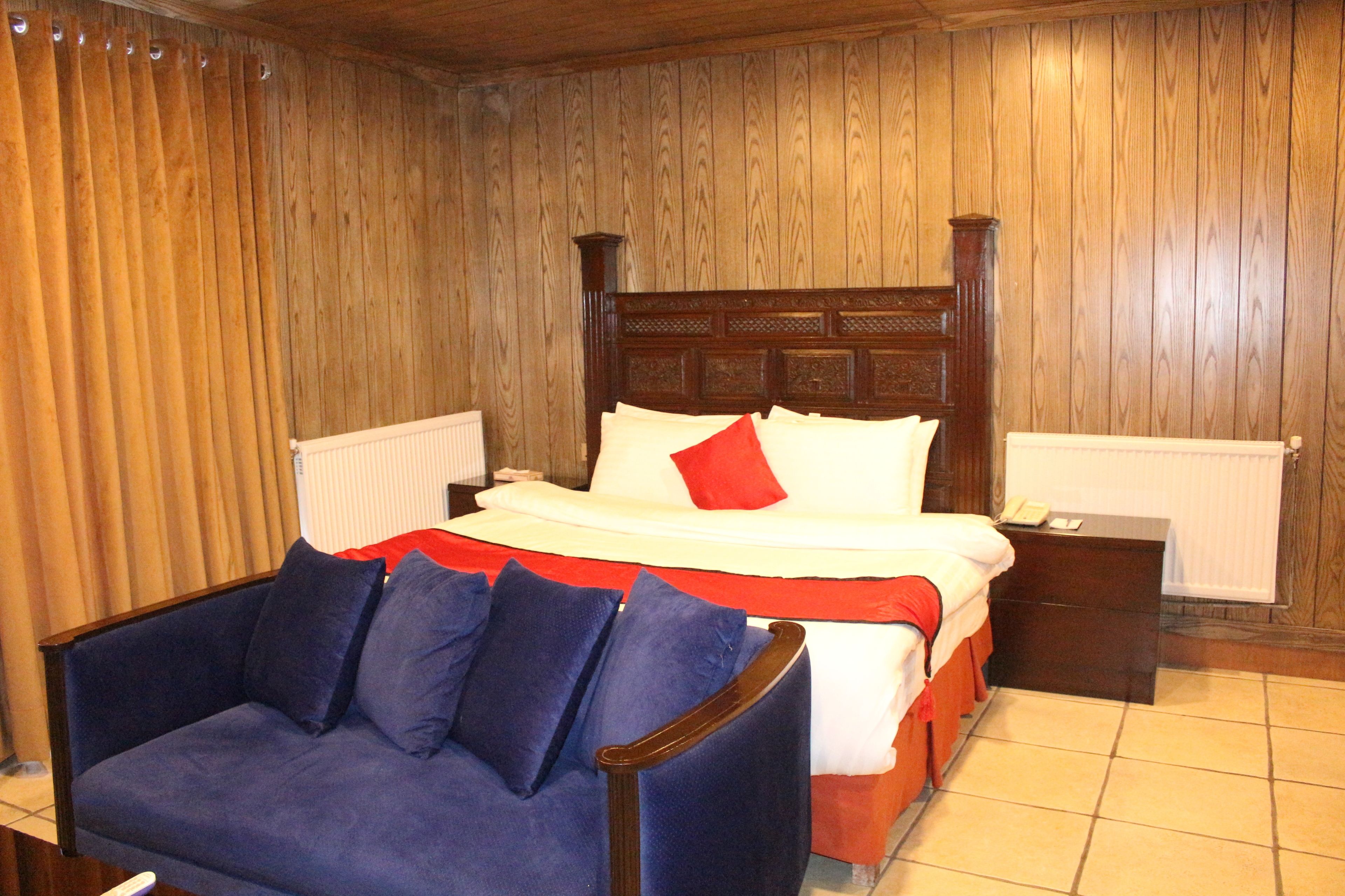 Un dormitorio con pared de paneles de madera, una cama con ropa de cama blanca y un cojín rojo, un sillón azul y una mesita de noche con un teléfono.