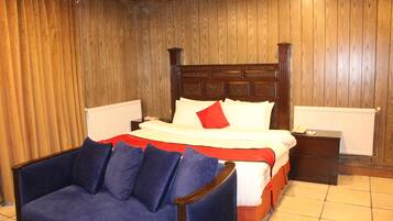 Un dormitorio con pared de paneles de madera, una cama con ropa de cama blanca y un cojín rojo, un sillón azul y una mesita de noche con un teléfono.