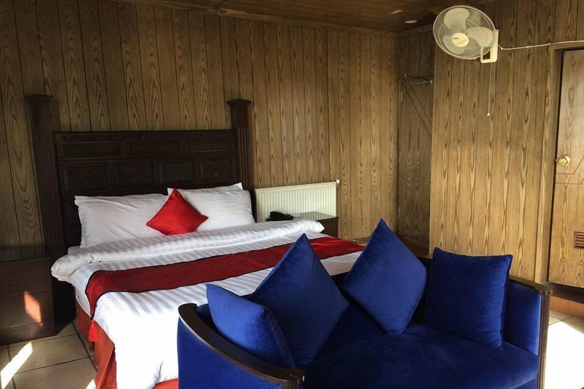 Un dormitorio con cabecera de madera, cama con ropa de cama blanca y roja, sofá azul y un ventilador de pie.