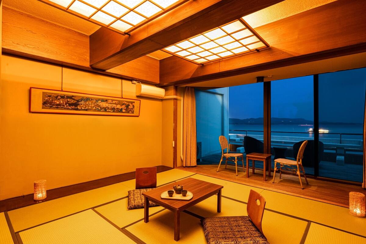 Une pièce de style japonais traditionnel, avec un sol recouvert de tatamis, une petite table sur laquelle est disposé un service à thé, et une vue sur la mer grâce à de grandes fenêtres.