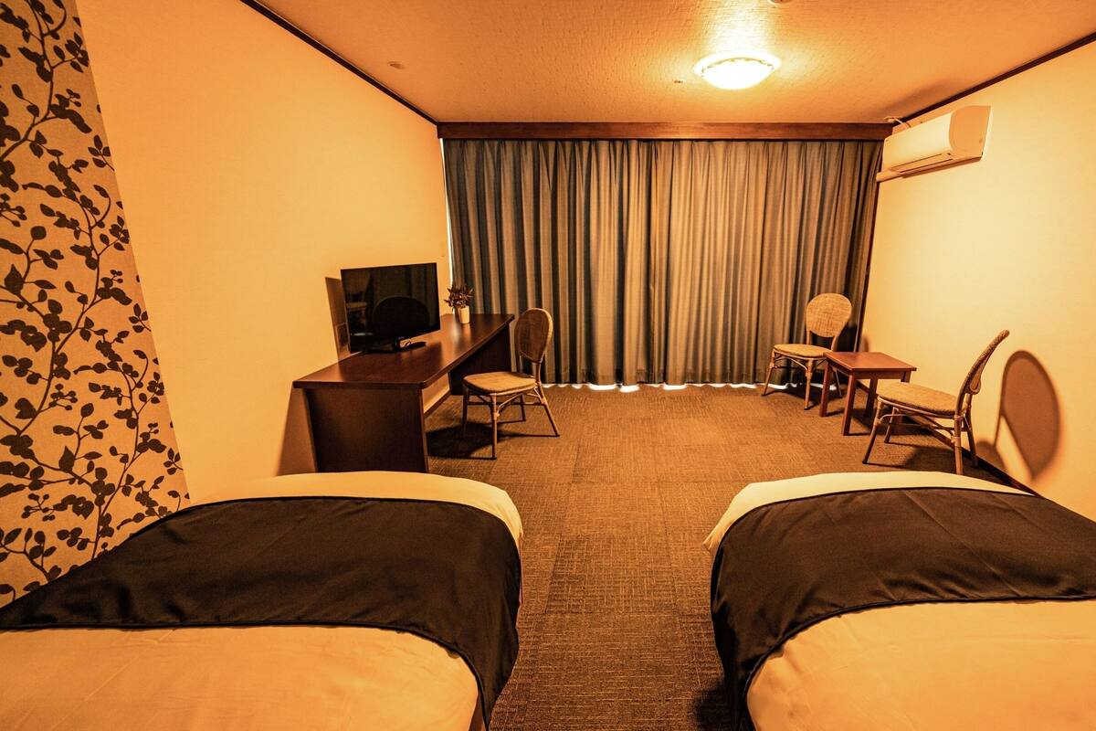 Une chambre d’hôtel avec deux lits, un bureau avec une télévision, une chaise, une petite table et une fenêtre avec des rideaux.