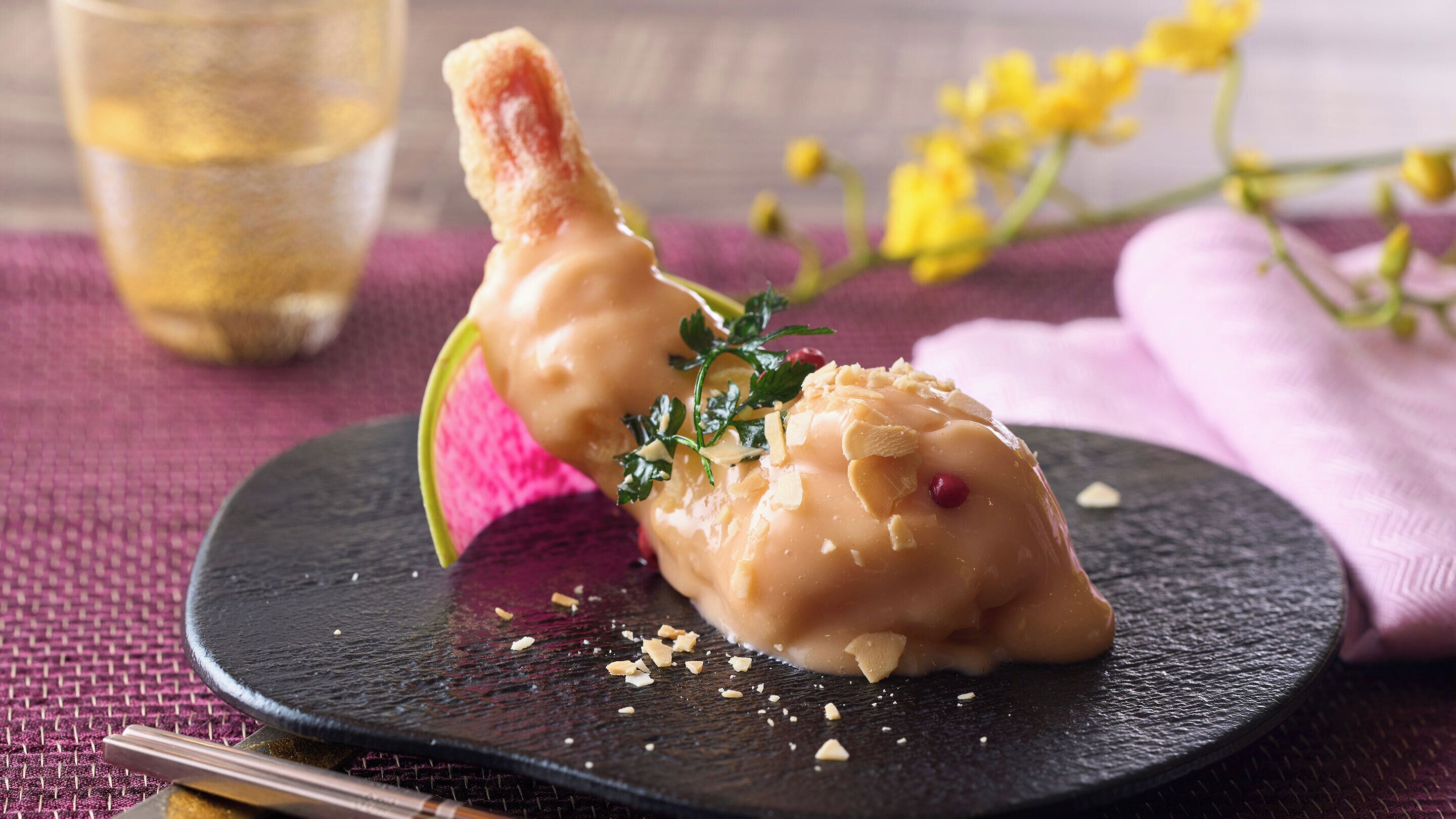 A dessert on a black plate with a fork, garnished with herbs and nuts, on a textured surface with a pink cloth and yellow flowers in the background.