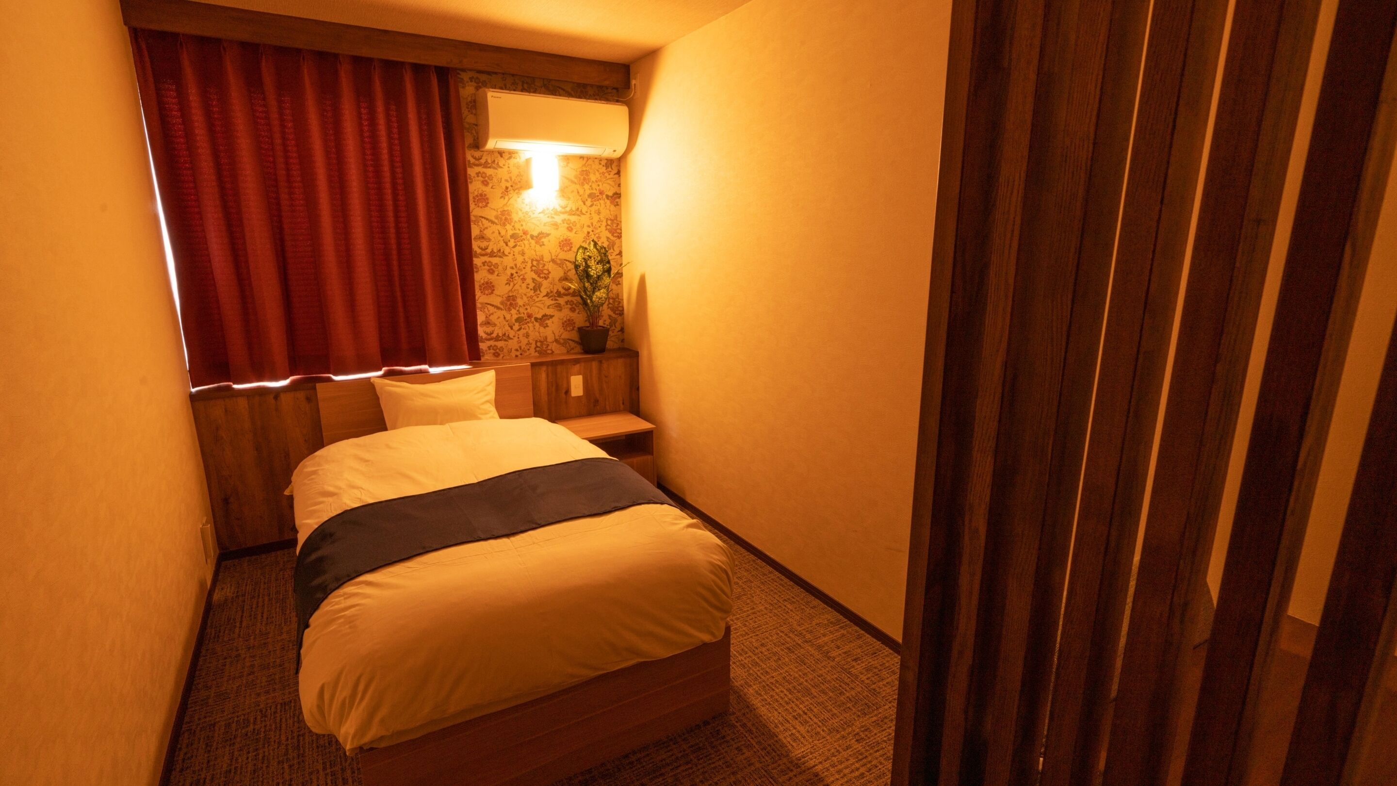 A hotel room with a bed, a nightstand, a potted plant, and a window with curtains.