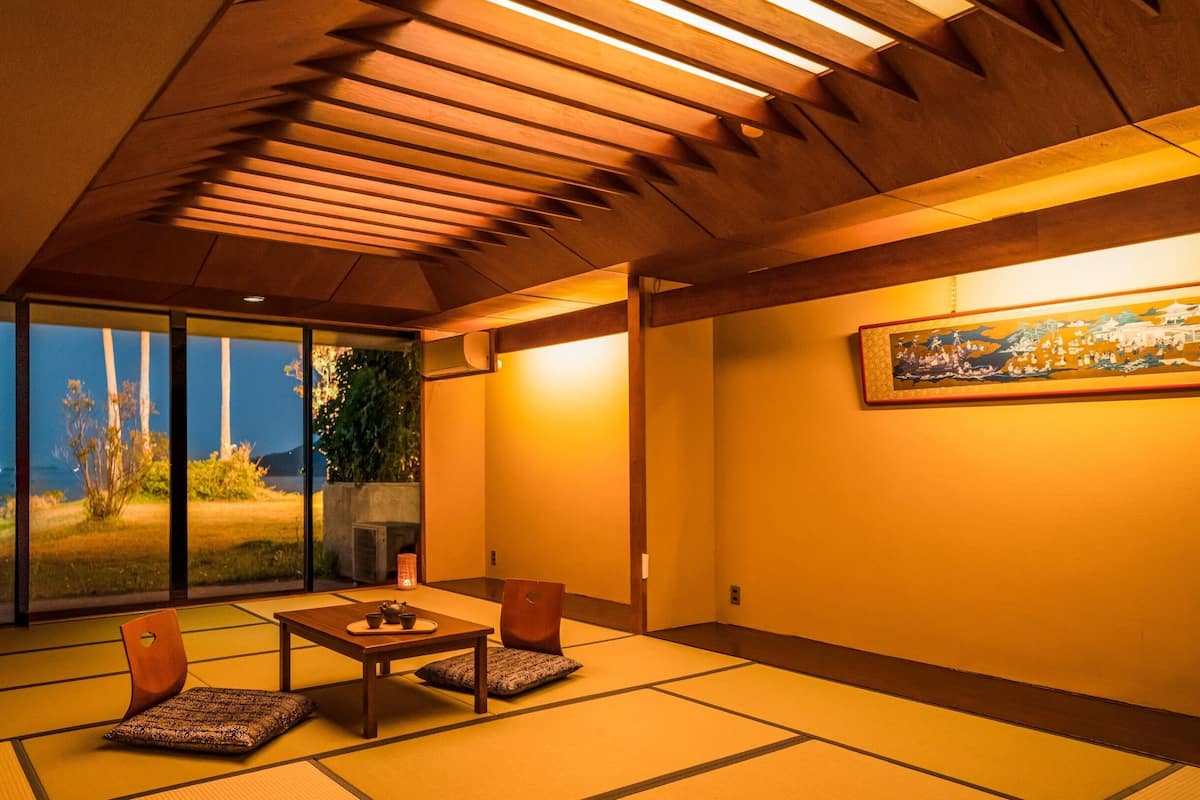 Une pièce de style japonais traditionnel, avec un sol recouvert de tatamis, une porte coulissante en verre donnant sur un jardin, une petite table avec un service à thé et un tableau accroché au mur.