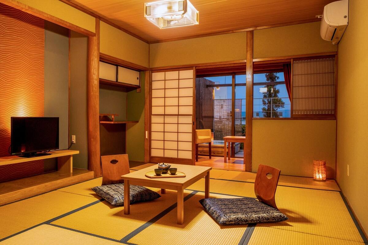 Une pièce traditionnelle japonaise avec un sol recouvert de tatamis, une table basse, un téléviseur posé sur une étagère et des portes coulissantes en papier de riz (shoji).