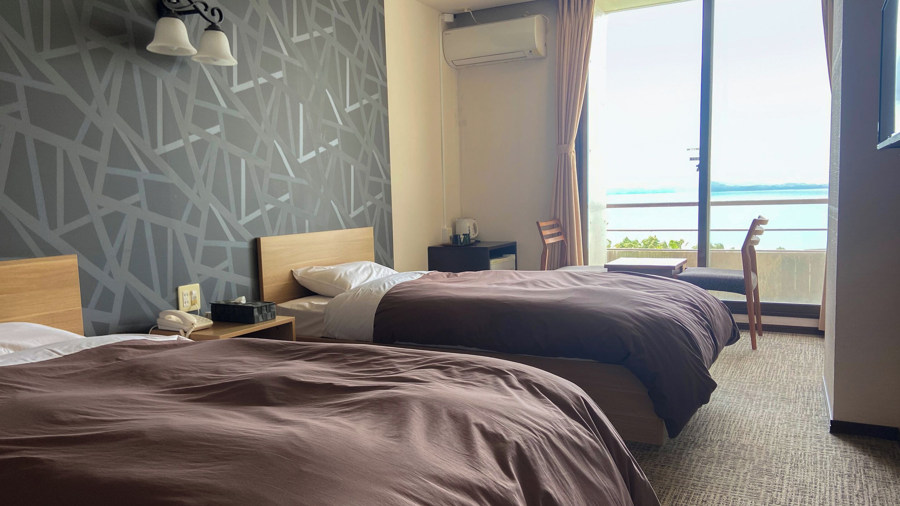 Ein Hotelzimmer mit zwei Betten, einem Nachttisch, einem Sessel, einem Fenster mit Meerblick und einer Wand mit geometrischem Muster.