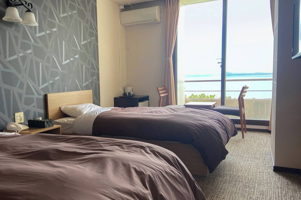Une chambre d’hôtel avec deux lits, une table de chevet, une chaise, une fenêtre donnant sur la mer et un mur orné d’un motif géométrique.