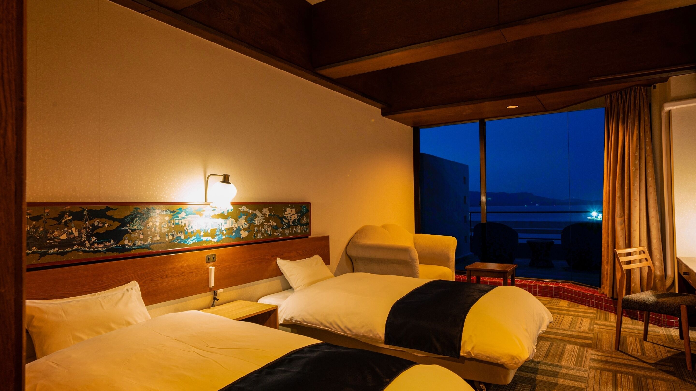 Ein Hotelzimmer mit einem großen Bett, einem Schreibtisch, einem Stuhl und Blick auf die beleuchtete Stadt.