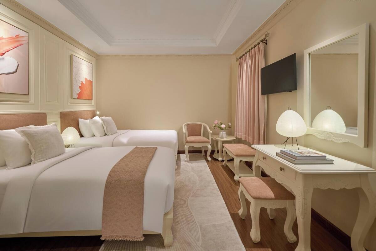 A hotel room with two beds, a desk with a lamp, a chair, a small table with a vase of flowers, and a large mirror.