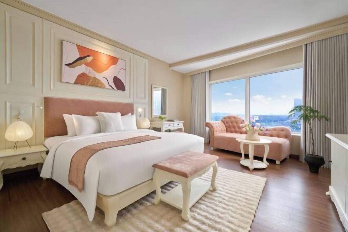 A hotel room with a large bed, a small bench, a round table, a sofa, and a large window offering a city view.