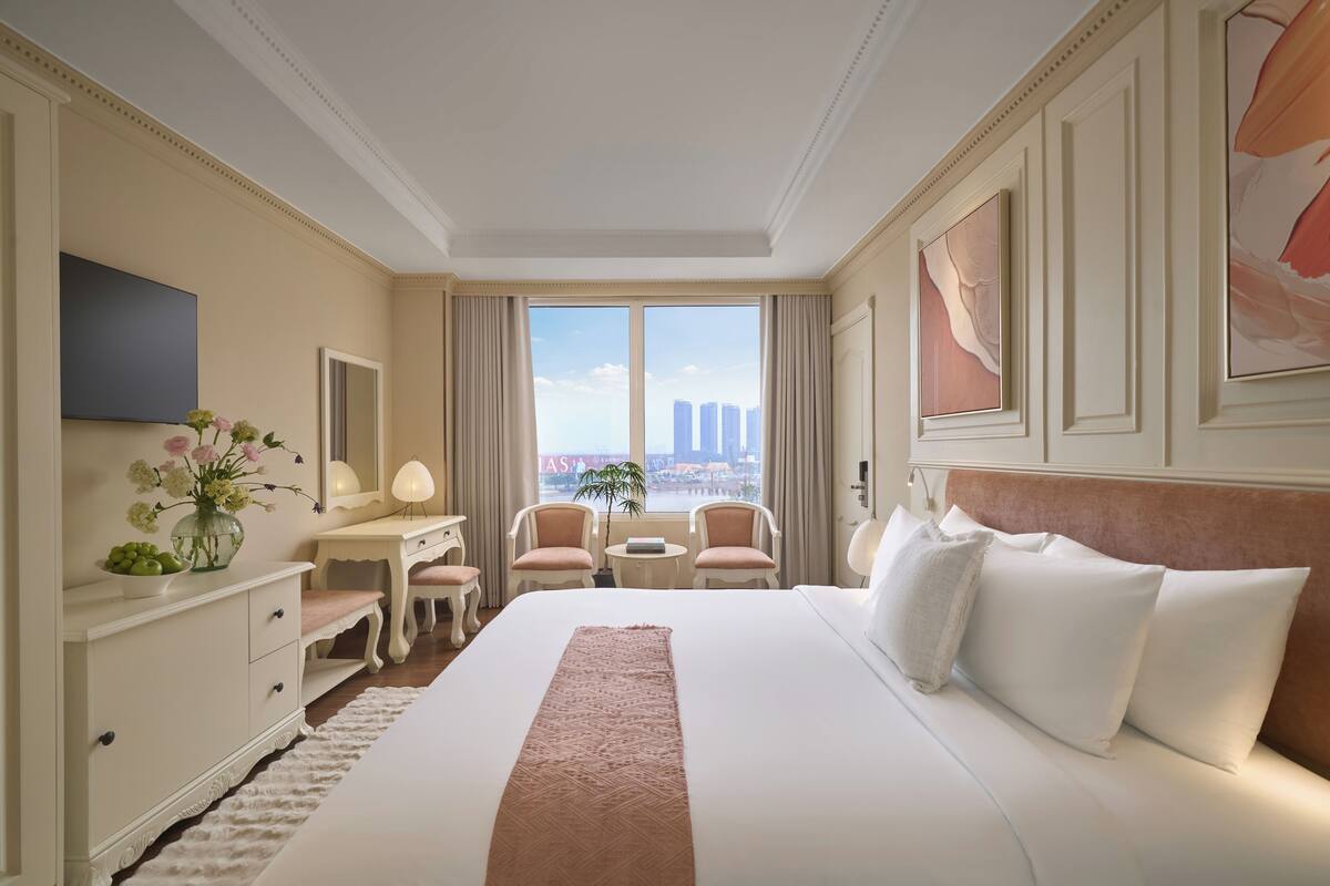 A spacious hotel room with a large bed, a desk, two chairs, a small table, and a view of tall buildings.