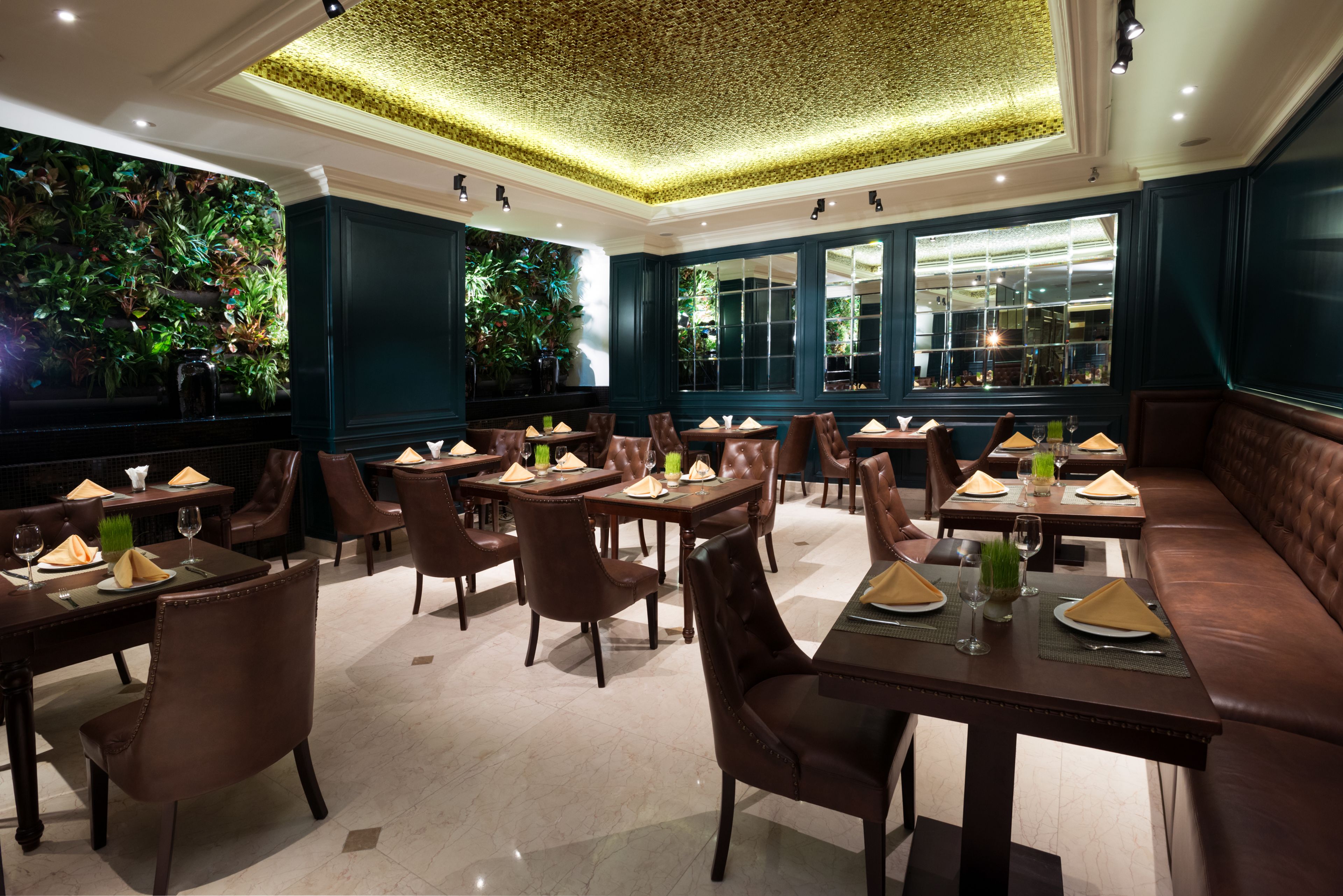 A restaurant with dark green walls, large windows, and a ceiling with a textured pattern. Tables are set with brown chairs, green napkins, and wine glasses.