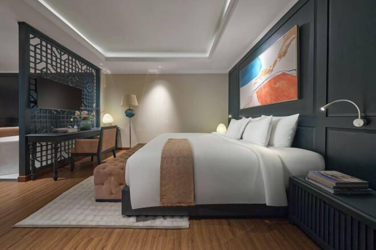 A modern hotel room with a bed, a desk, a chair, and a large abstract painting on the wall.