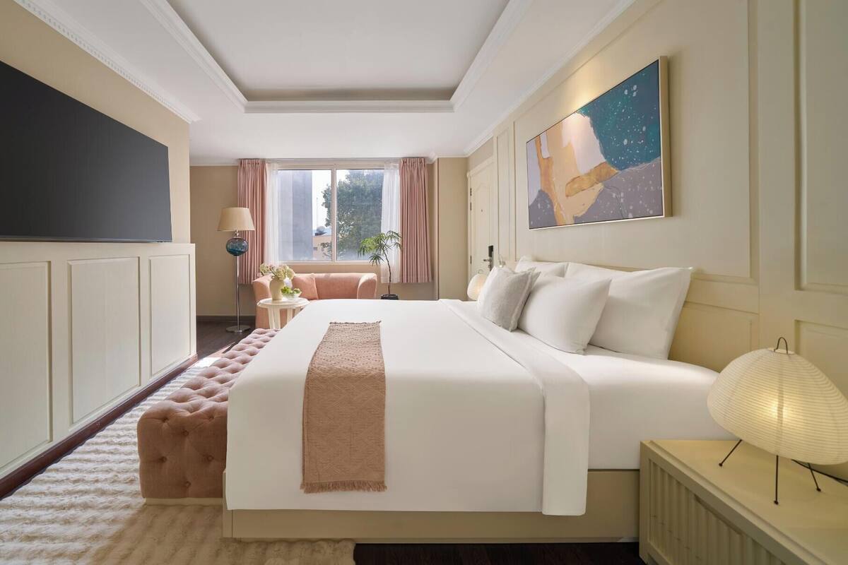 A hotel room with a large bed, a flat-screen TV, a desk with a lamp, and a window with curtains.