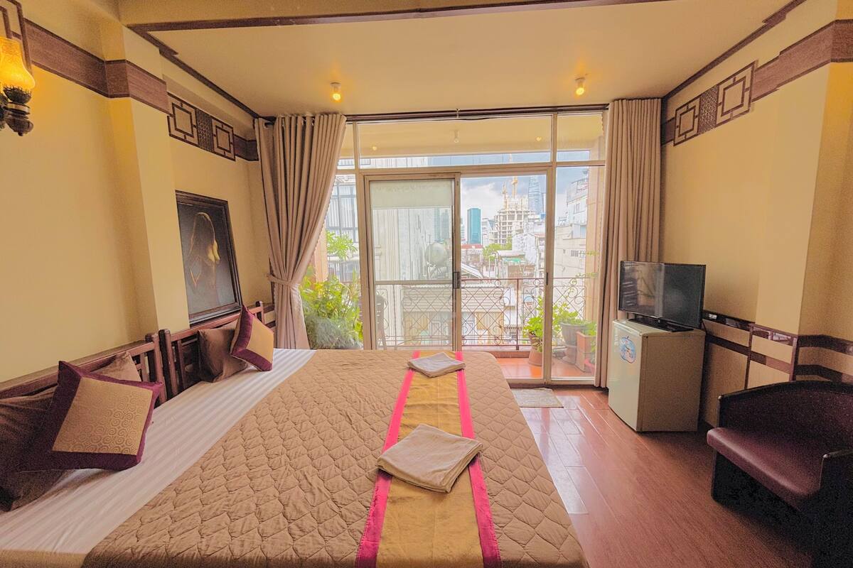 A hotel room with a large bed, a TV, a desk, and a balcony with a city view.