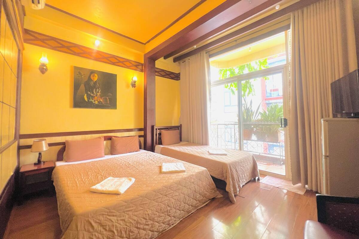 A hotel room with two beds, a painting on the wall, a balcony with curtains, and a television.