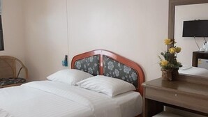 Un lit bien fait, avec du linge de lit blanc, une tête de lit en bois ornée de motifs floraux, une table de chevet sur laquelle est posé un vase de fleurs jaunes, et un téléviseur à écran plat fixé au mur.