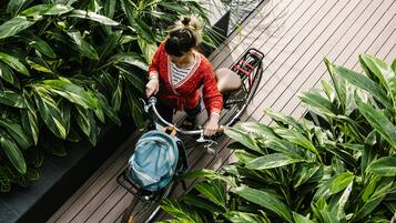A person riding a bicycle with a blue bag on a wooden deck surrounded by green plants.