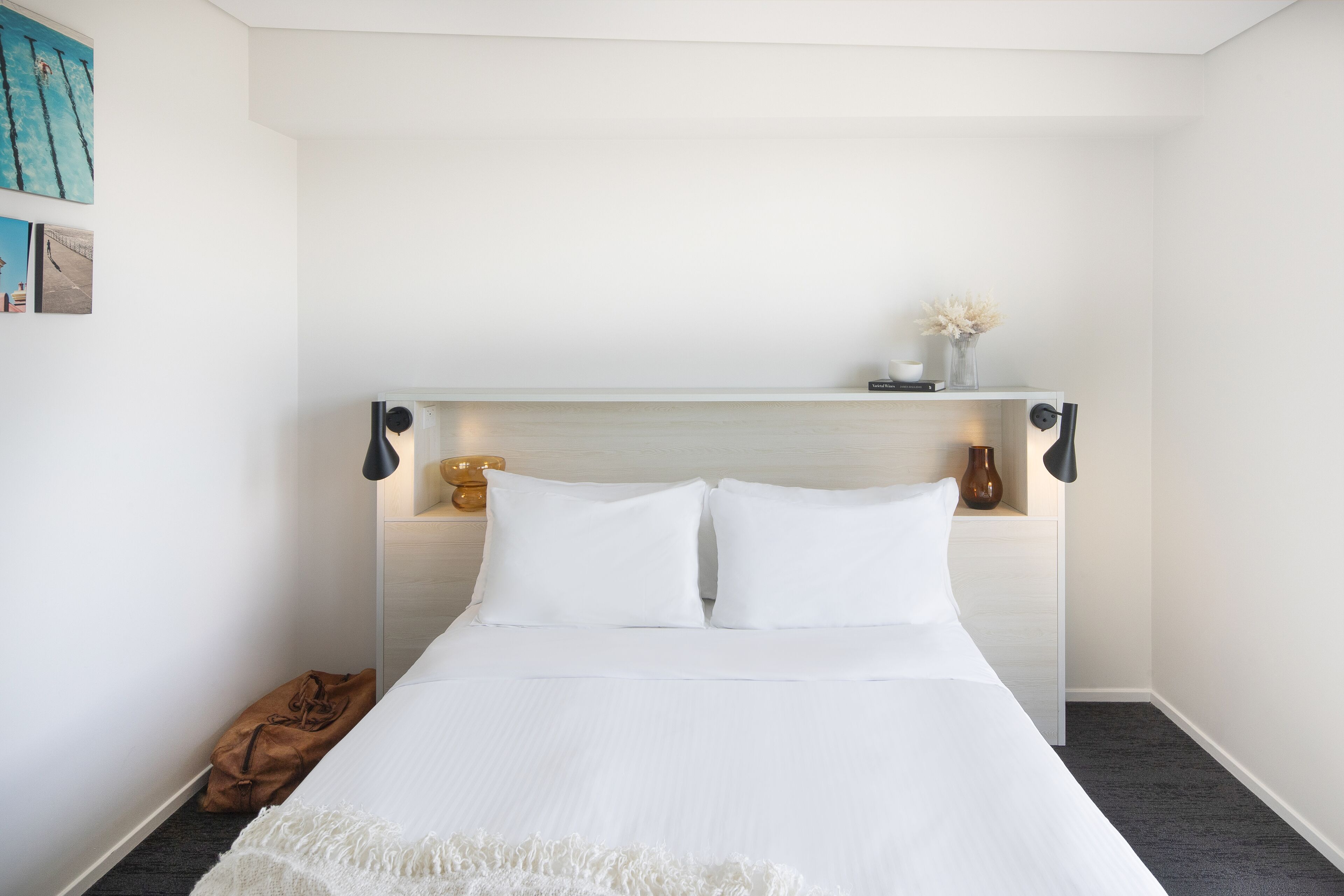 A neatly made bed with white linens, a brown throw blanket, and a small brown bag on the floor.