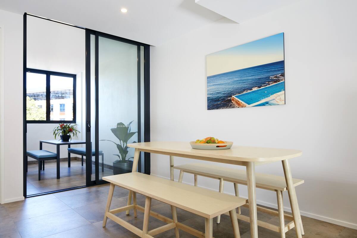 A modern dining area with a wooden table and bench, a potted plant, and a view of the sea through a large window.