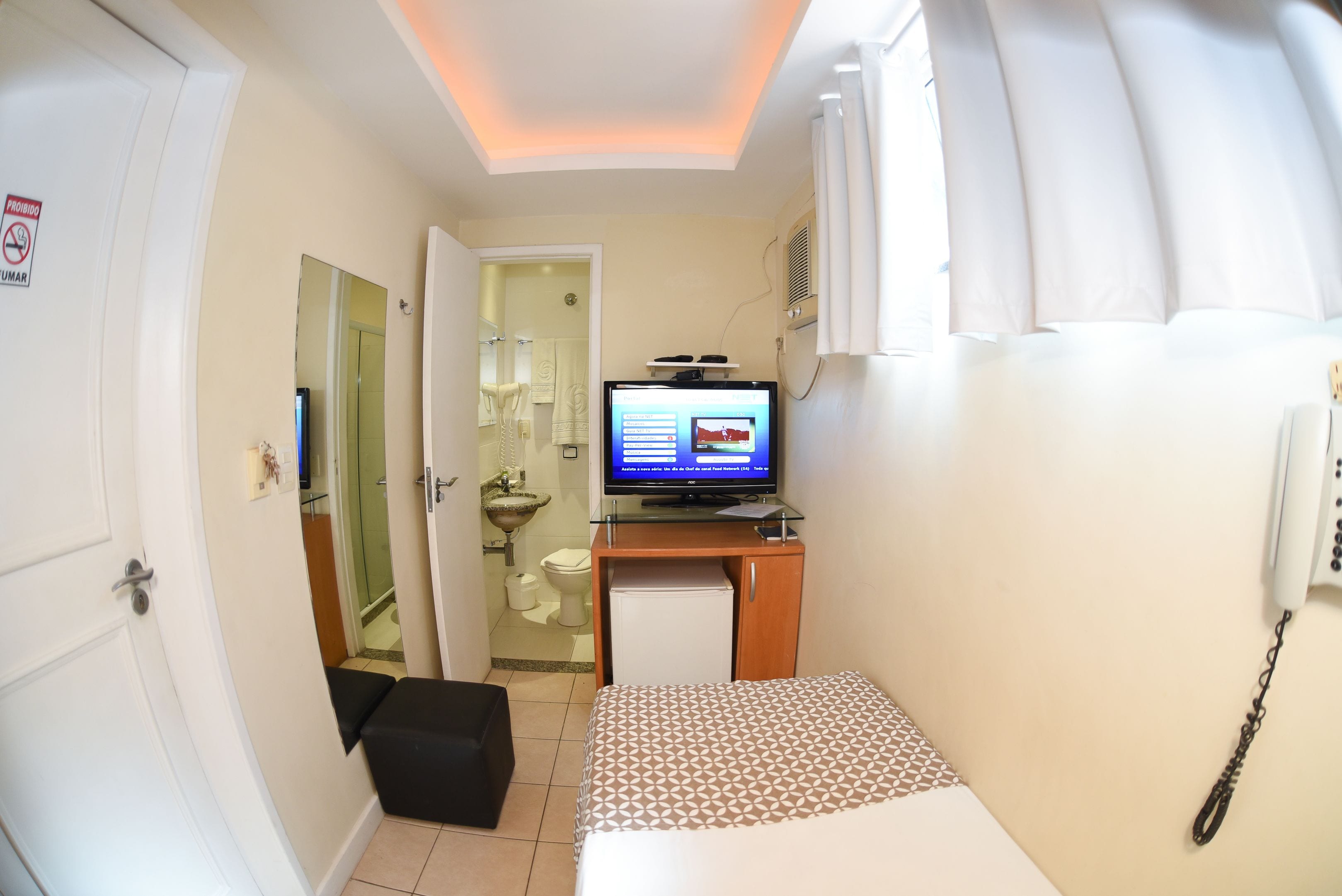 A compact hotel room with a bed, a small desk with a television, a mini-fridge, and a bathroom visible through an open door.